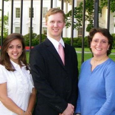 A man and two women stand outside a fence around a white building, all dressed in business attire